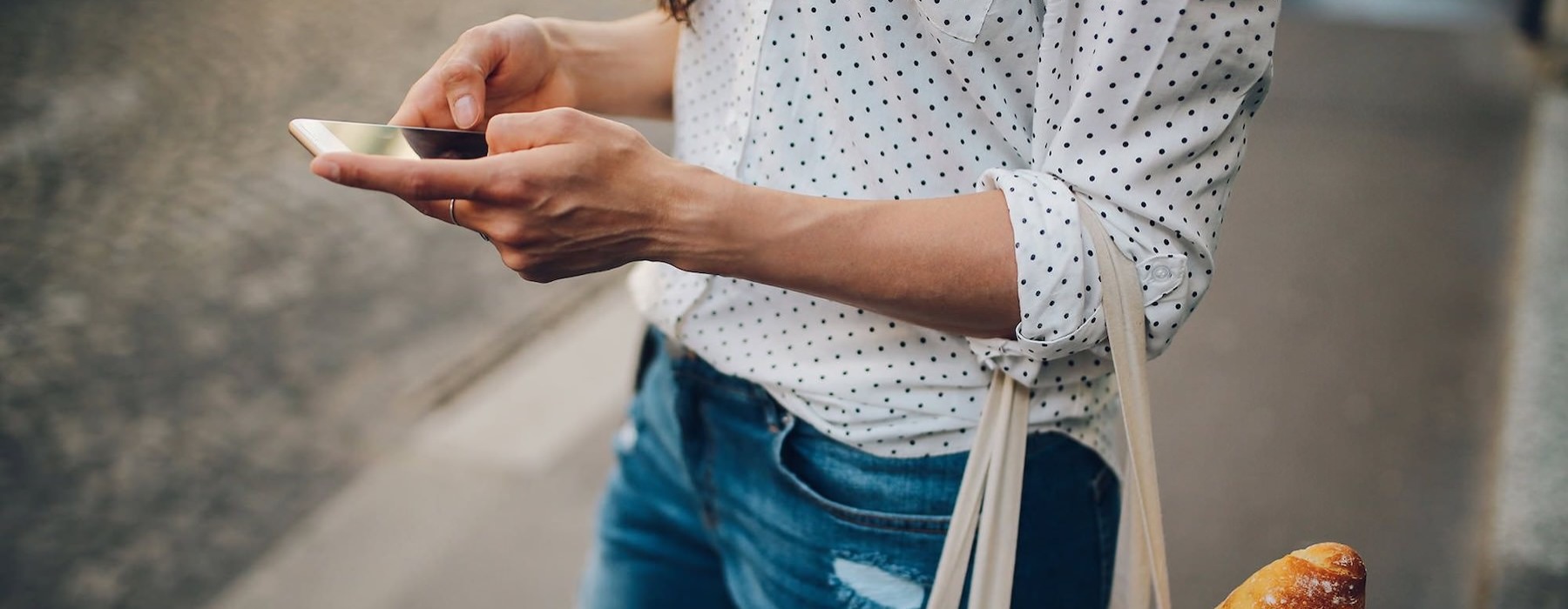 woman walks down the street and texts with a bag of groceries on her arm